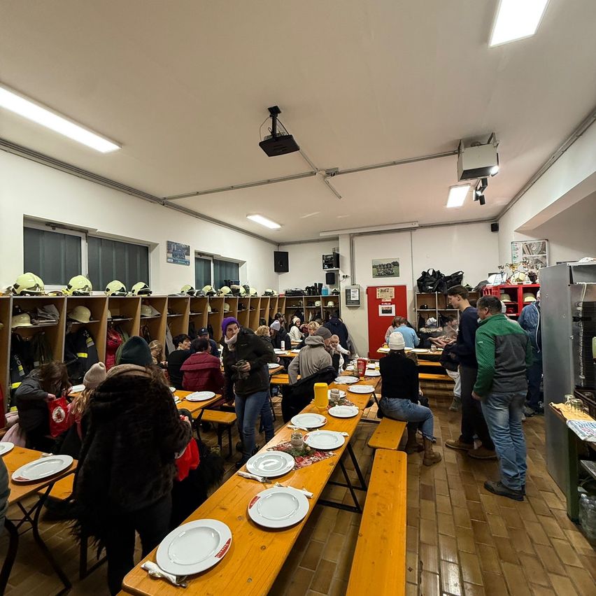 Eine Gruppe von Menschen ist drinnen versammelt, an langen Holztischen sitzend. Die Tische sind mit Tellern beladen und der Raum ist mit gelben Helmen an Regalen geschmückt. Einige Personen stehen in der Nähe der Tische und unterhalten sich.