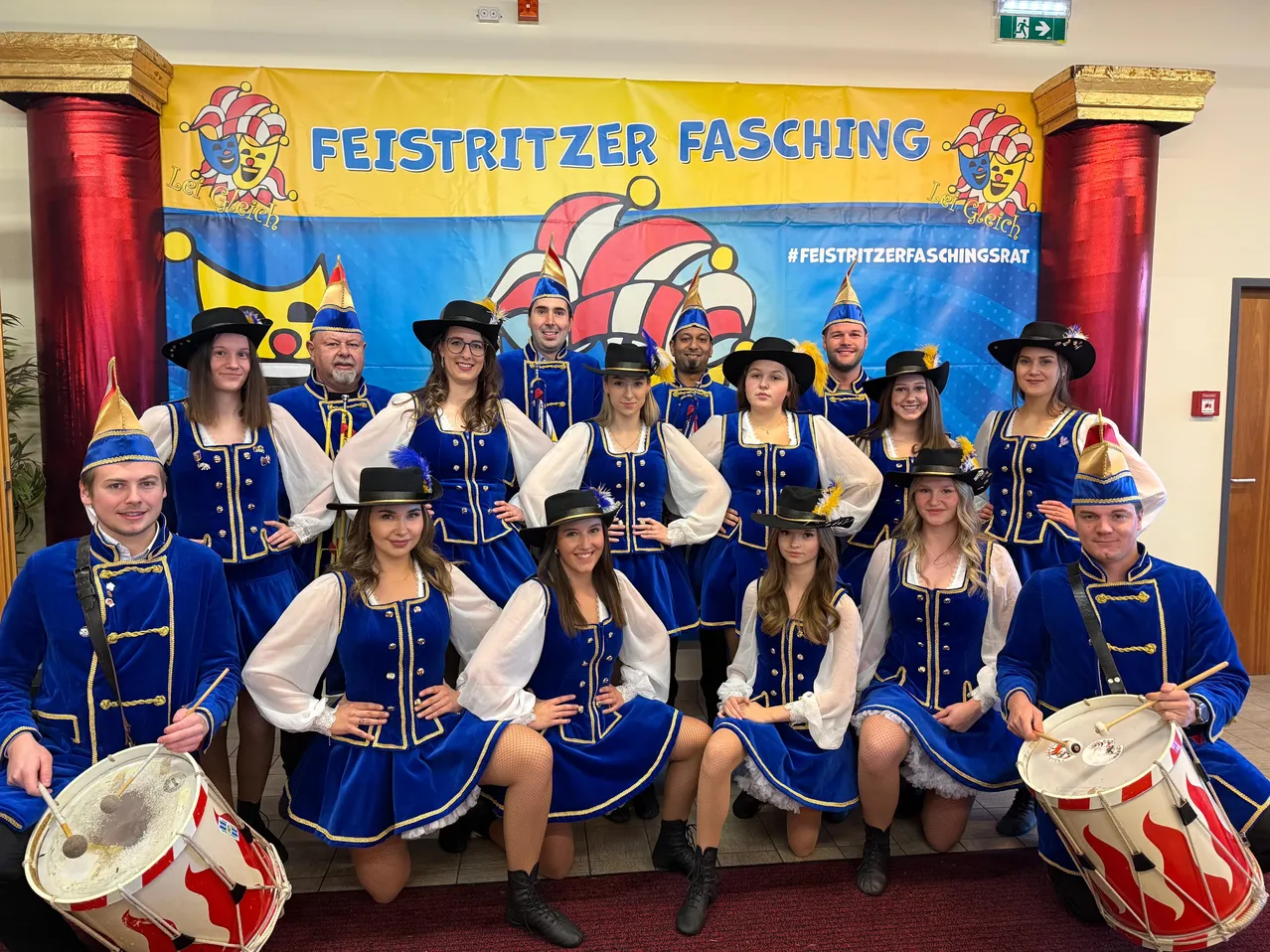 Eine Gruppe von Menschen in traditionellen blauen Kostümen mit goldenen Akzenten und Hüten posiert für ein Foto vor einem gelben Banner mit den Worten 'Feistritzer Fasching'. Sie halten Trommeln und lächeln.