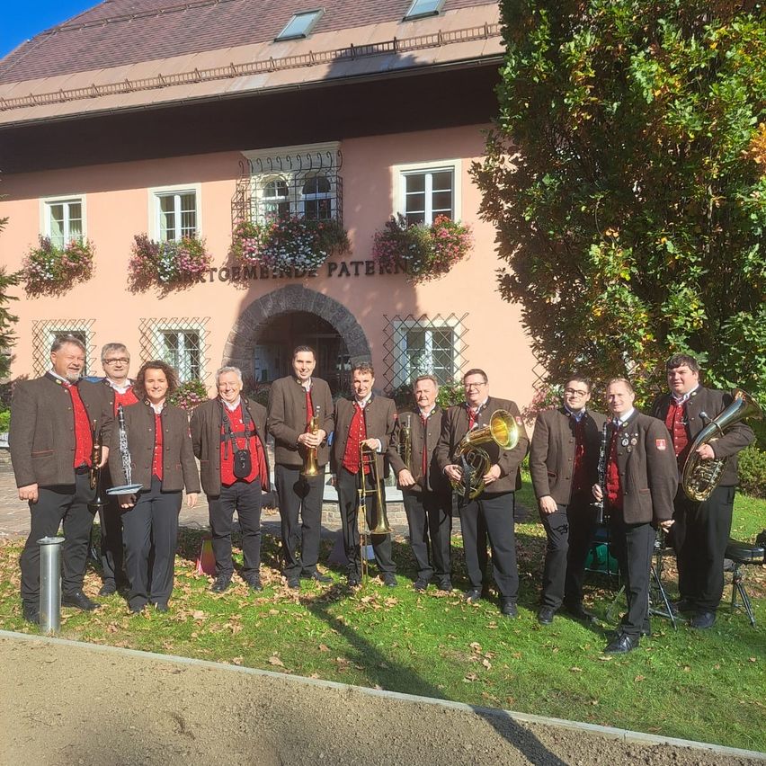 Eine Gruppe von Musikern steht vor einem rosa Gebäude mit einem Baum und Herbstblättern. Das Gebäude trägt den Text 'STORMBUDE PATERN'.