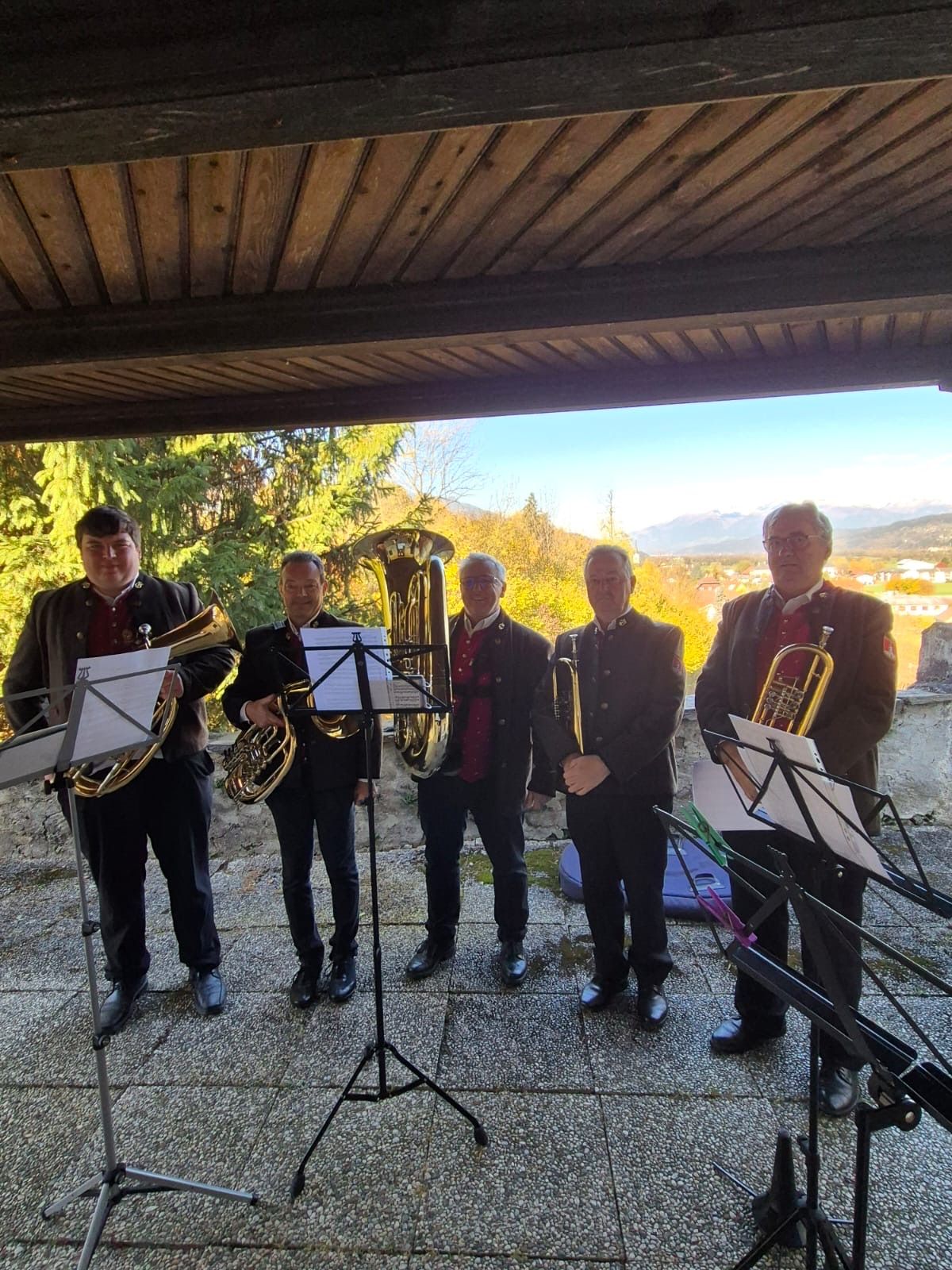 Fünf Männer in Uniformen stehen unter einem Pavillon, halten Blechblasinstrumente und scheinen sich auf eine Aufführung vorzubereiten. Hinter ihnen sind Bäume und Berge in der Ferne.