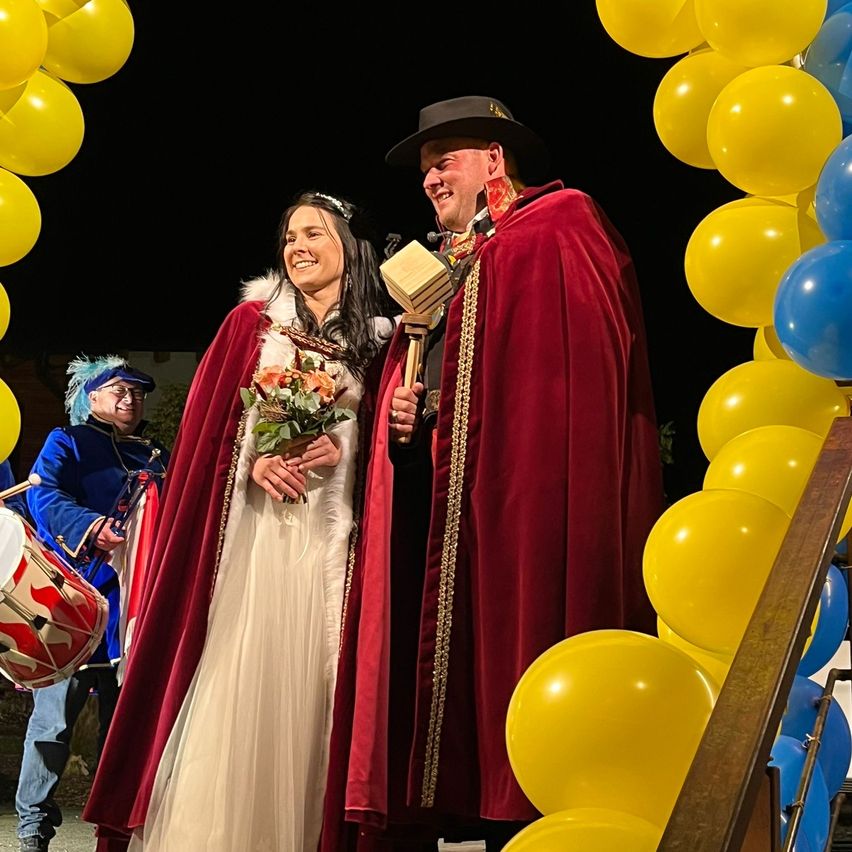 Ein lächelndes Paar in roten Umhängen steht bei Nacht unter einem Ballonbogen. Ein Mann hält ein Buch und ein Mikrofon, während eine Frau einen Blumenstrauß hält. Ein Mann in einer blauen Uniform spielt hinter ihnen eine Trommel.