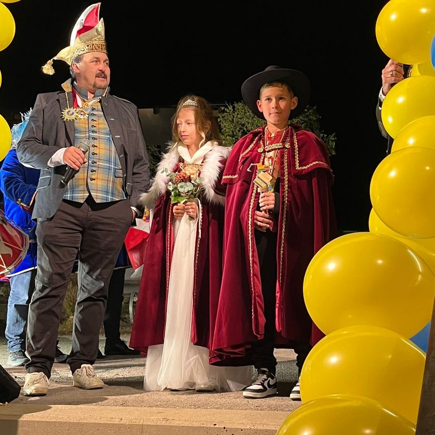 Ein Mann in Kostüm hält ein Mikrofon und steht neben einem jungen Mädchen in einem weißen Kleid und einem Jungen in einem roten Umhang, umgeben von gelben Luftballons, vermutlich bei einer Veranstaltung.