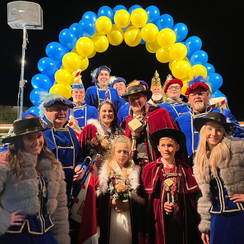 Eine Gruppe von Menschen in blauen und roten Kostümen und Hüten posiert für ein Foto vor einem blauen und gelben Luftballonbogen. Einige halten Trophäen und Blumen.