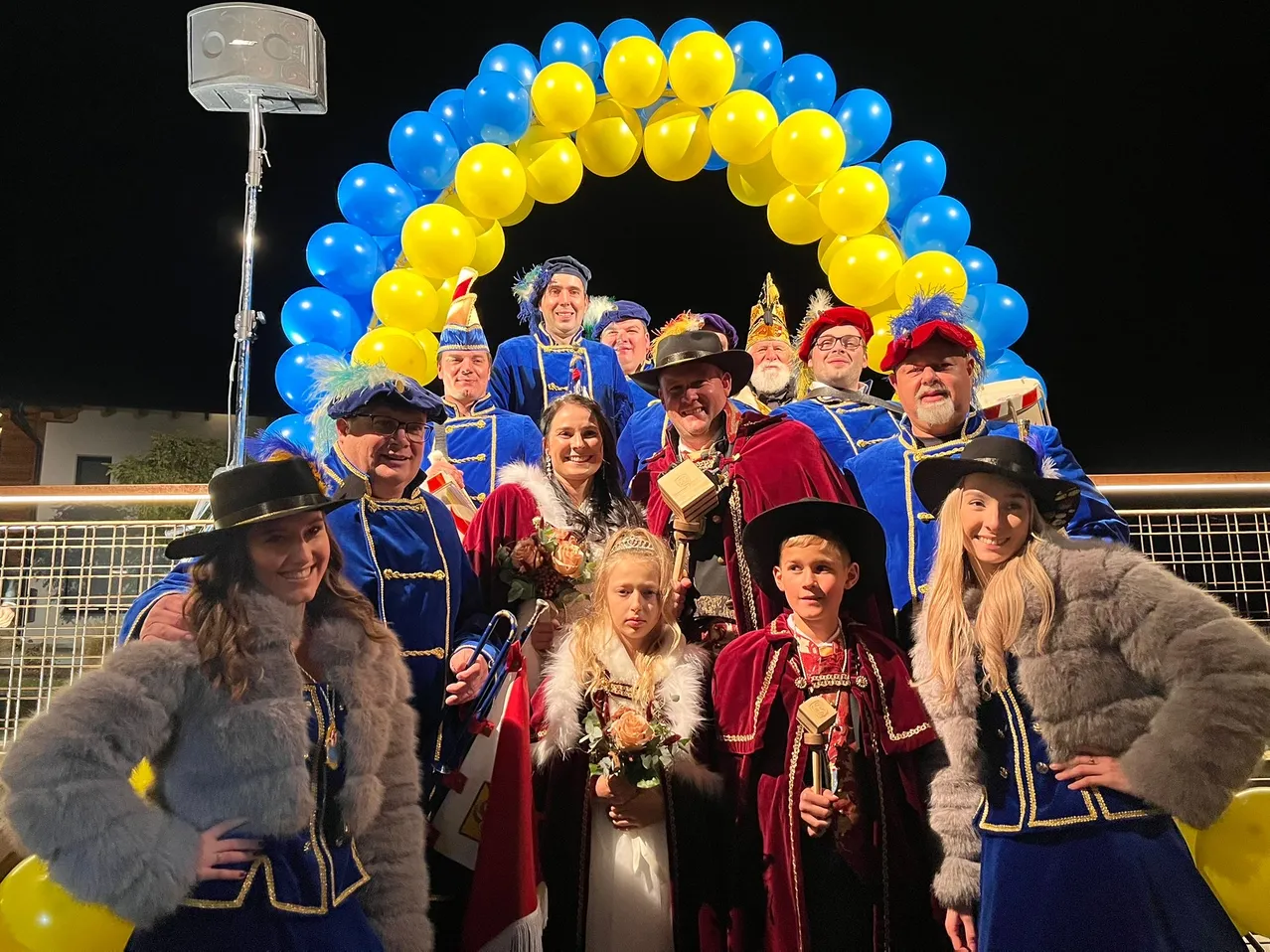 Eine Gruppe von Menschen in blauen und roten Kostümen und Hüten posiert für ein Foto vor einem blauen und gelben Luftballonbogen. Einige halten Trophäen und Blumen.