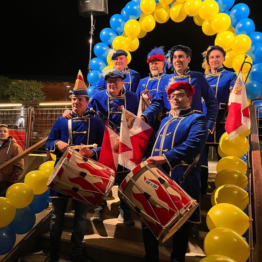 Eine Gruppe von Männern in blauen Uniformen und Hüten spielt auf Trommeln und steht auf einer Treppe mit Luftballons. Einige halten Fahnen und haben einen Lautsprecher hinter sich.