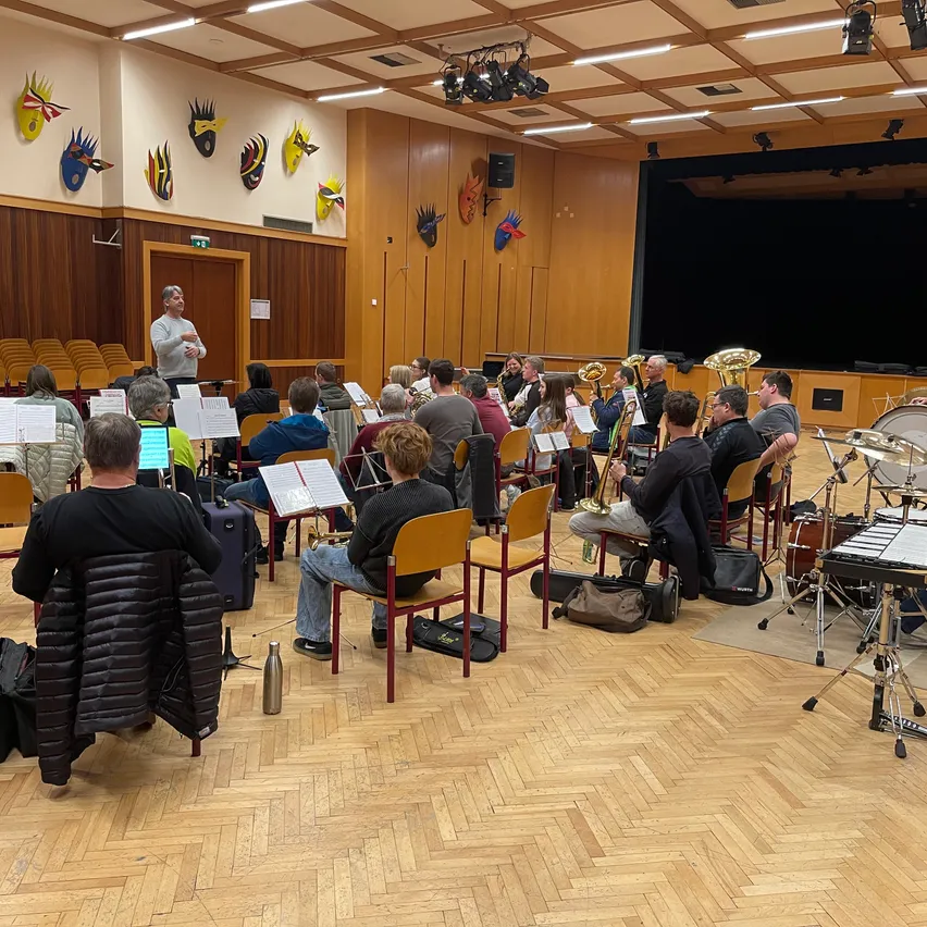 Eine Gruppe von Menschen sitzt in Stühlen und spielt Instrumente. Ein Mann steht vor ihnen und dirigiert. Notenblätter liegen auf ihren Knien. Der Raum hat Holzböden und Wände.