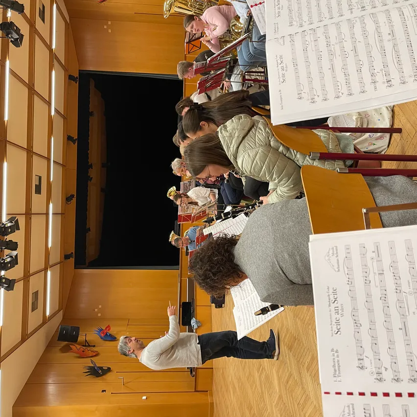 Eine Gruppe von Musikern ist in einem Saal versammelt und übt für eine Aufführung. Der Dirigent steht in der Mitte und deutet, während andere Instrumente spielen. Notenblätter sind auf Stühlen um sie herum ausgebreitet.