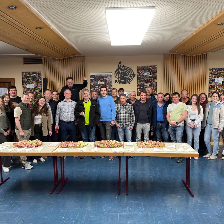 Eine Gruppe von Menschen posiert für ein Foto in einem Raum mit gedeckten Tischen, einige lächeln und ein Mann steht auf einem Tisch.