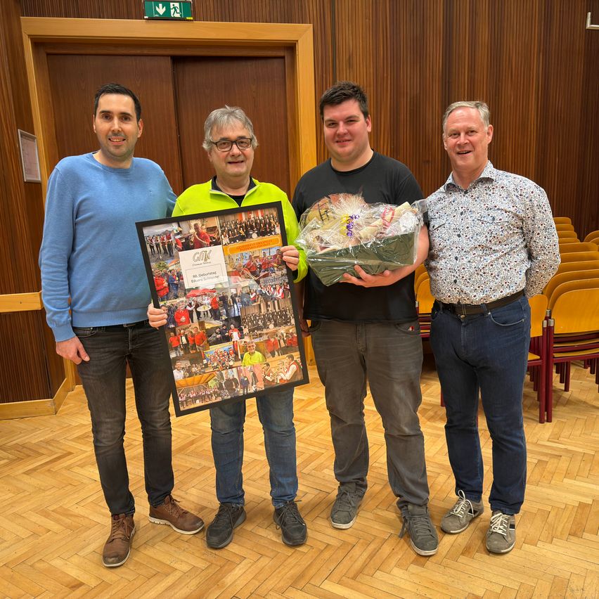 Vier Männer stehen lächelnd in einem Raum zusammen und halten ein gerahmtes Foto und einen Blumenstrauß. Der Mann auf der linken Seite trägt einen blauen Pullover, während der Mann in der Mitte einen Strauß hält.