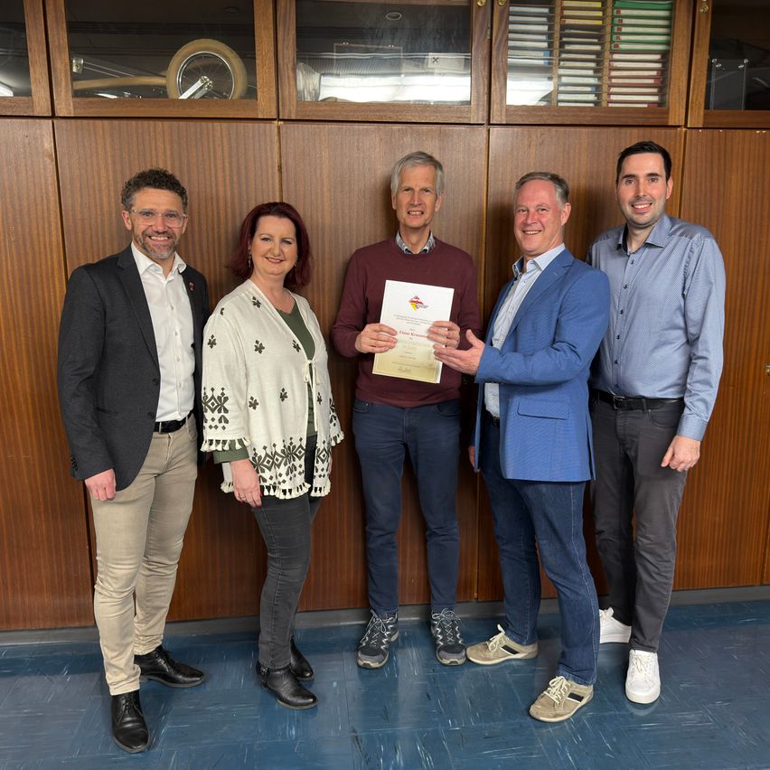 Fünf Personen stehen vor einem hölzernen Hintergrund und lächeln für ein Foto. Ein Mann hält ein Zertifikat.