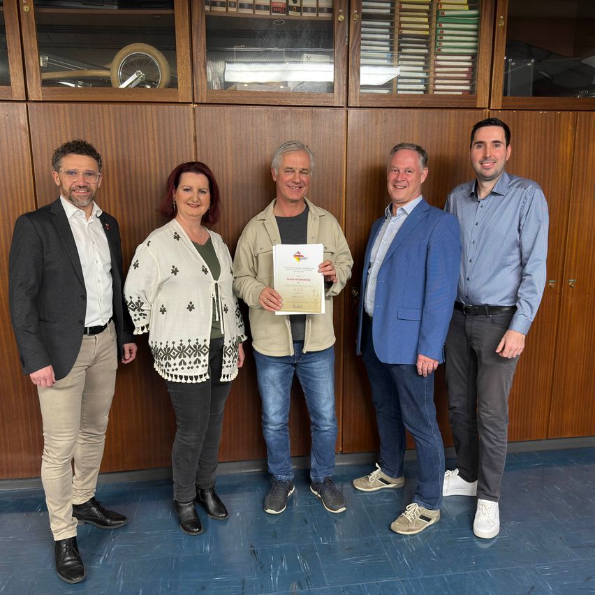 Fünf Menschen stehen lächelnd vor Holzschränken. Der Mann in der Mitte hält ein Zertifikat. Der Mann rechts trägt ein blaues Hemd und einen Gürtel. Die Frau links trägt einen weißen Schal. Der Mann links trägt einen schwarzen Mantel.