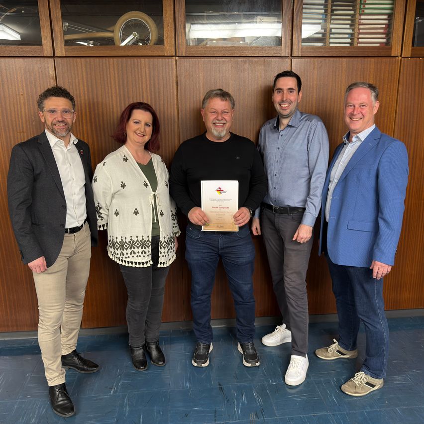 Fünf Erwachsene stehen in einem Raum und lächeln für ein Foto. Ein Mann hält eine Urkunde. Hinter ihnen eine Holzwand mit Regalen und Lampen.