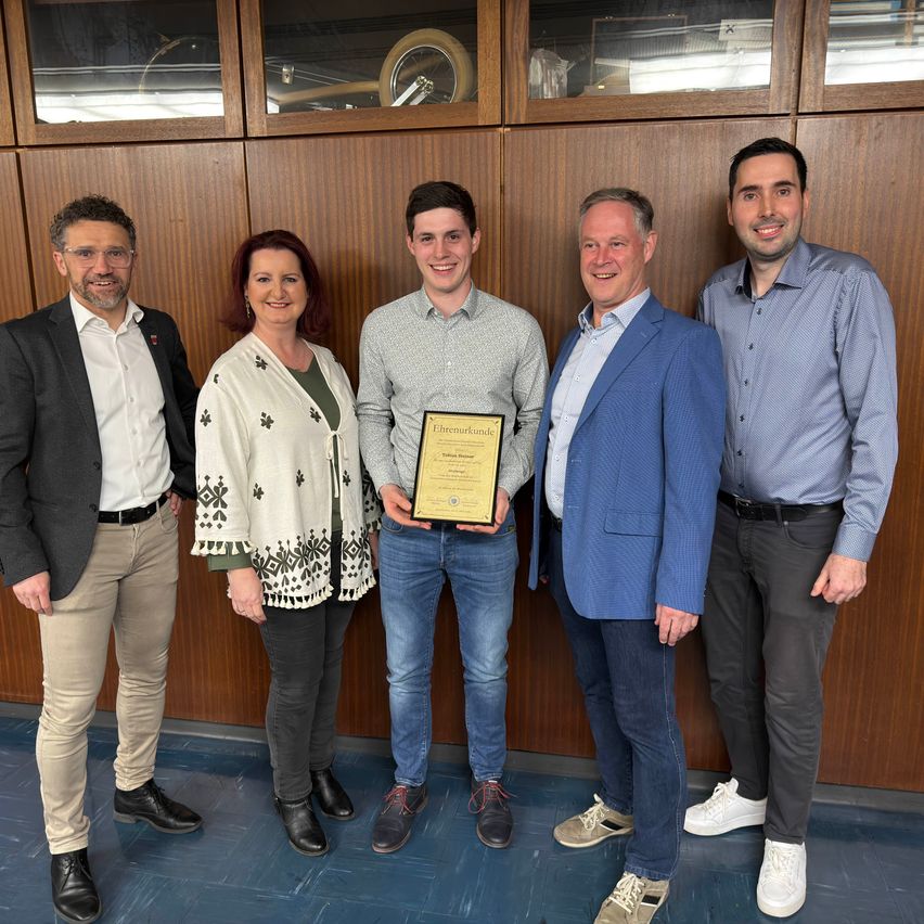 Fünf Menschen stehen lächelnd in einem Raum zusammen. Der Mann in der Mitte hält ein Zertifikat. Sie stehen vor einer Holzwand mit Regalen und einer Glasvitrine.
