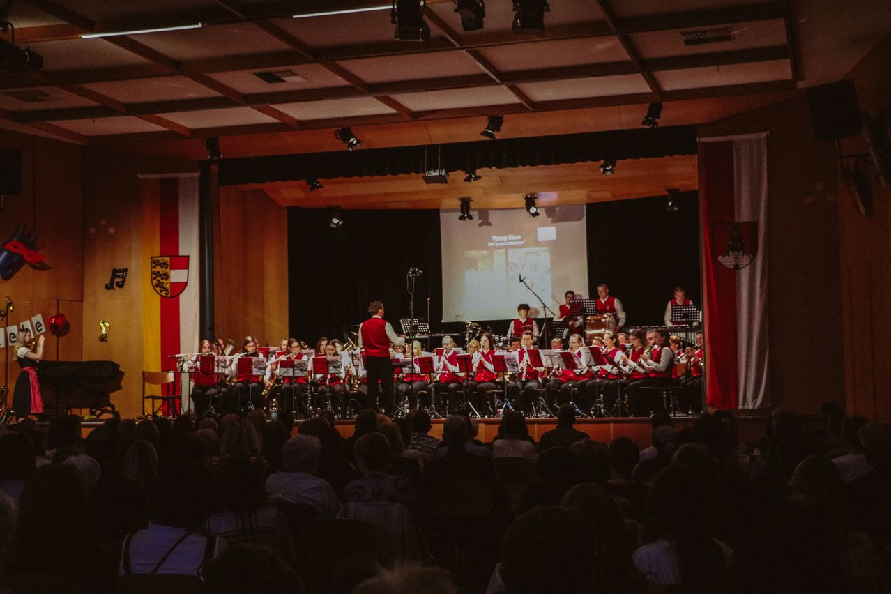 Eine Schulband spielt auf der Bühne, während ein großes Publikum zuschaut. Scheinwerfer und eine Projektionsleinwand sind sichtbar.
