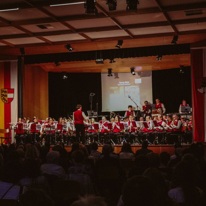 Eine Schulband spielt auf der Bühne, während ein großes Publikum zuschaut. Scheinwerfer und eine Projektionsleinwand sind sichtbar.
