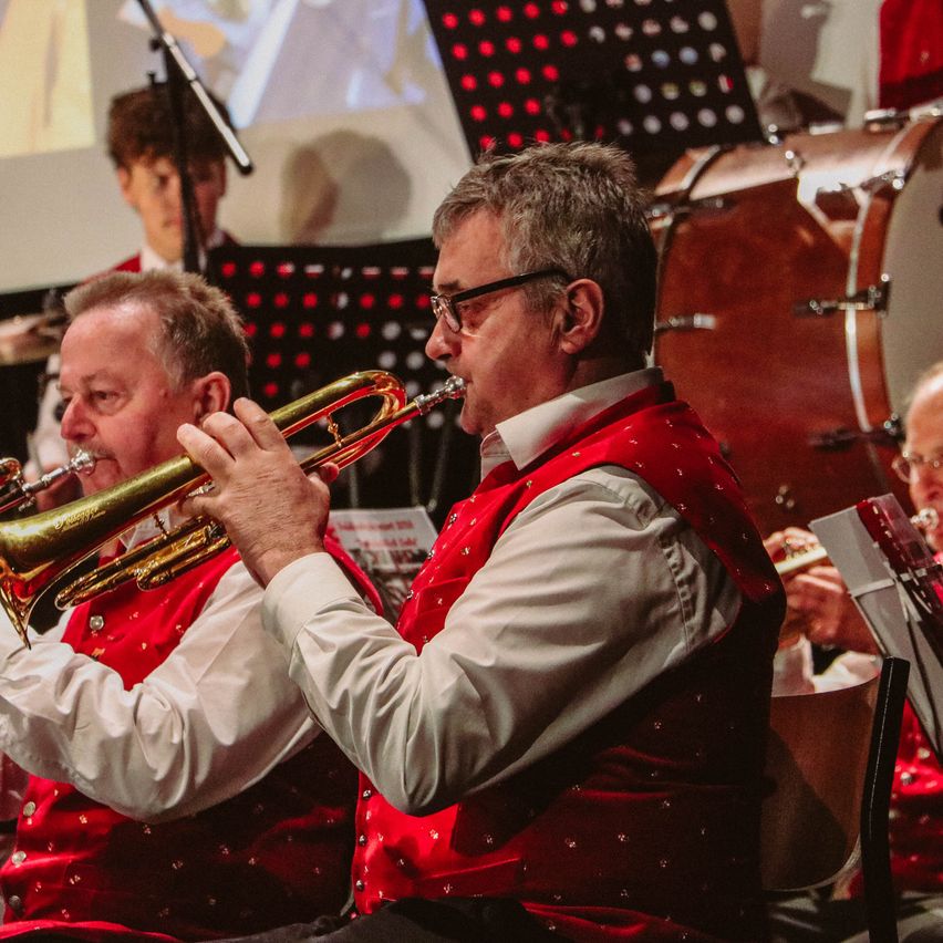 Eine Gruppe von Musikern in roten Westen spielt auf der Bühne, einer von ihnen spielt Trompete. Dahinter steht ein Schlagzeug und ein weiterer Musiker spielt Flöte.
