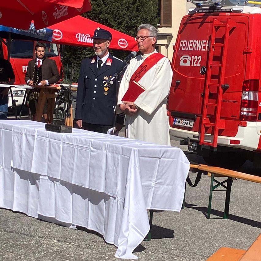 Ein Mann in einer weißen Robe steht neben einem Feuerwehrfahrzeug an einem Podium. Ein anderer Mann in blauer Uniform steht hinter ihm.