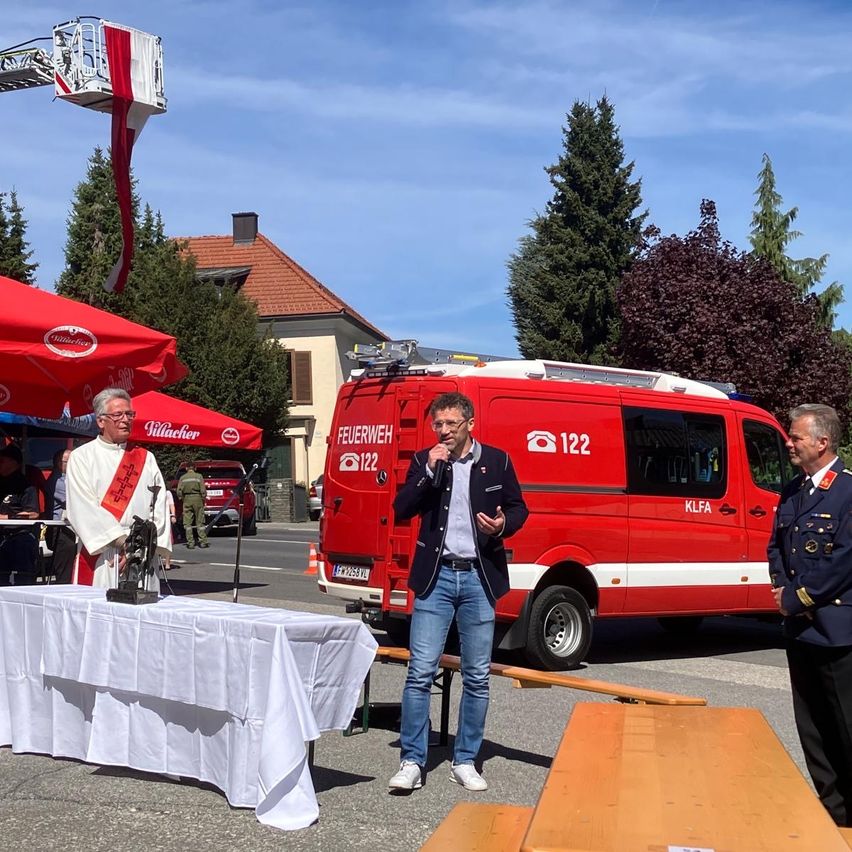 Ein Mann spricht in ein Mikrofon bei einer Veranstaltung im Freien. Er steht neben einem roten Feuerwehrwagen. Ein anderer Mann in Uniform steht rechts.