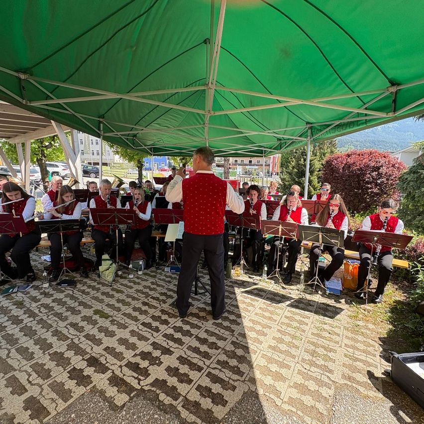 Ein Blechbläserensemble spielt unter einem grünen Zelt im Freien, mit Musikern in Reihen und einem Dirigenten vorne.