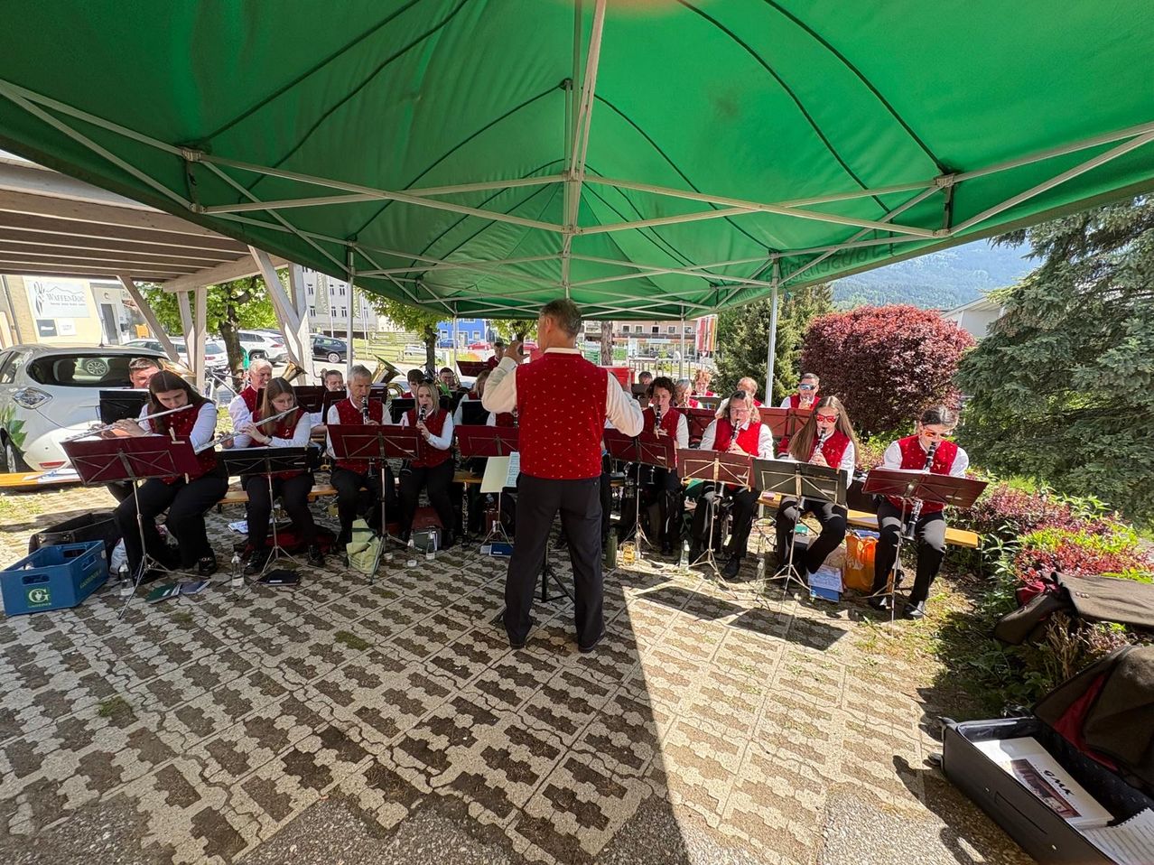 Ein Blechbläserensemble spielt unter einem grünen Zelt im Freien, mit Musikern in Reihen und einem Dirigenten vorne.
