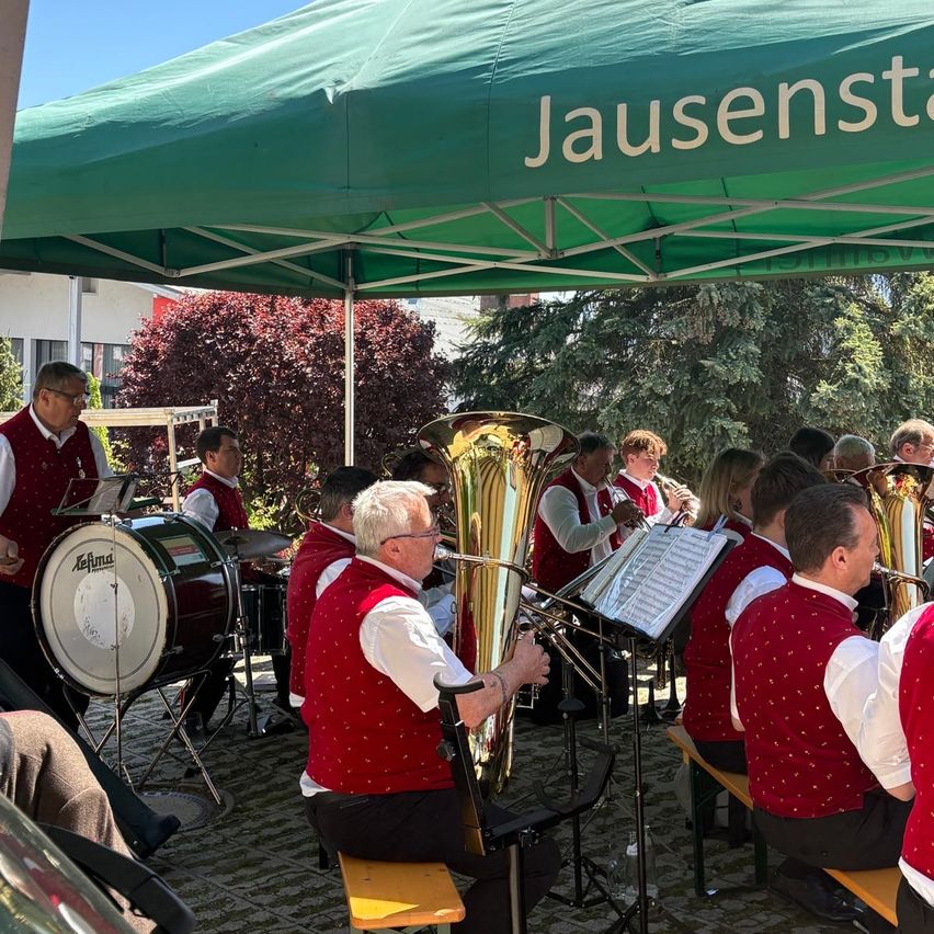 Ein Blasorchester spielt an einem sonnigen Tag unter einem grünen Zelt, mit Musikern auf Bänken, einer spielt Tuba und andere mit Trompeten und Posaunen.