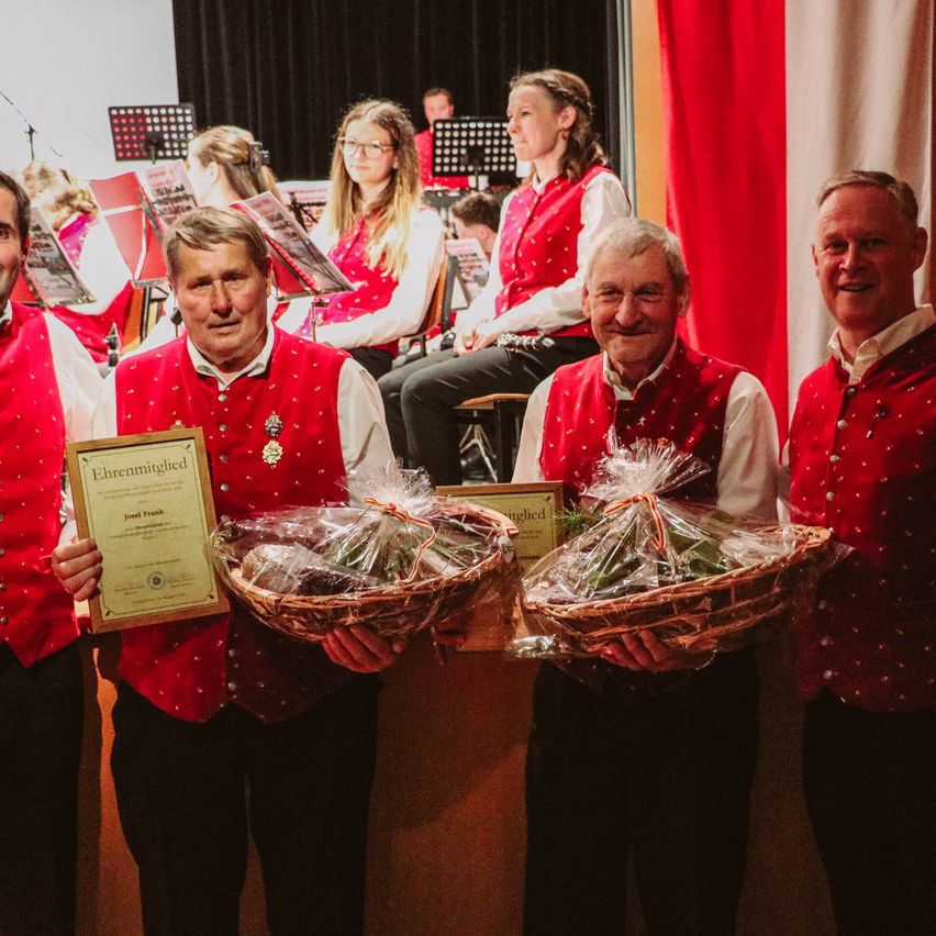 Vier Männer in roten Westen posieren mit Körben und einem Zertifikat. Hinter ihnen spielt eine Band in roten Westen Instrumente. Der Mann auf der linken Seite hält ein gerahmtes Zertifikat.