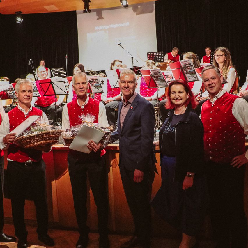 Fünf Menschen stehen vor einer Bühne mit einem Beamer und halten Körbe und Papiere. Dahinter steht eine Band, und Musiker sitzen auf Stühlen.