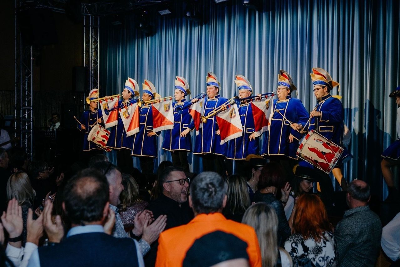 Eine Band in blauen Uniformen tritt mit Flaggen und Instrumenten vor einem Publikum auf der Bühne auf.
