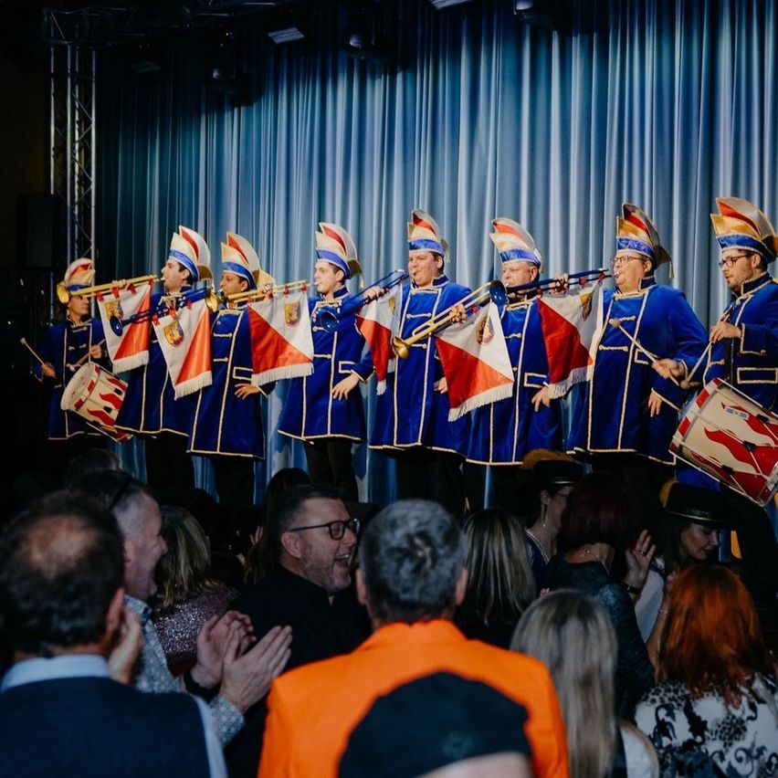 Eine Band in blauen Uniformen tritt mit Flaggen und Instrumenten vor einem Publikum auf der Bühne auf.