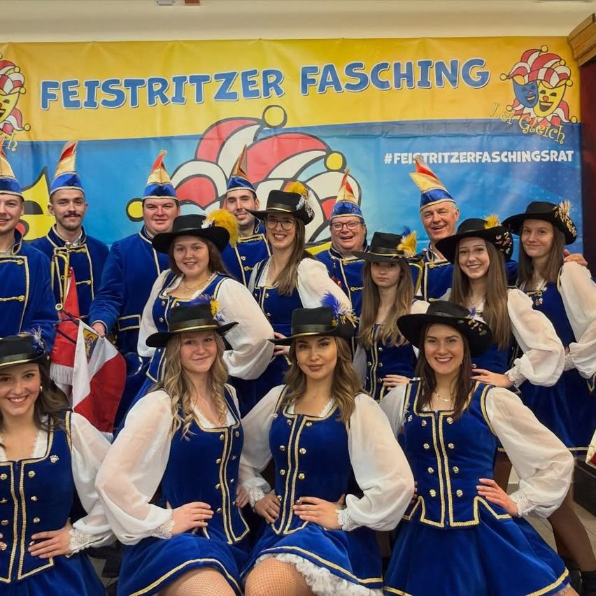 Eine Gruppe von Personen in blauen und weißen Kostümen und Hüten, die für ein Foto posieren. Ein Banner mit Text und einer Clown-Figur steht hinter ihnen.