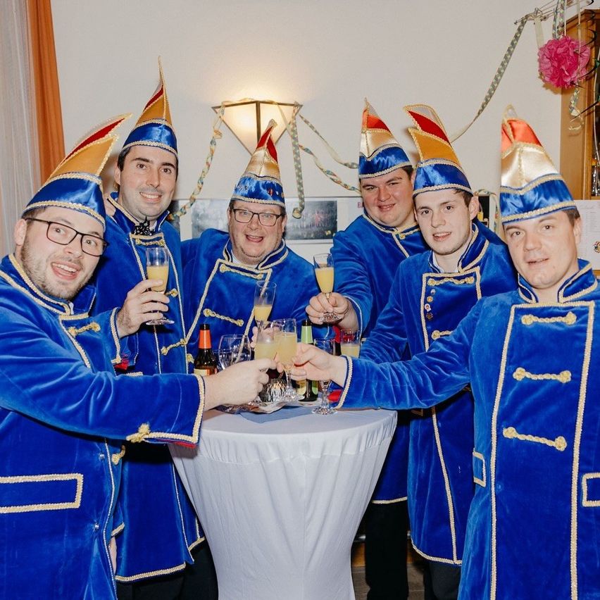 Eine Gruppe von Männern in blauen Kostümen mit goldener Einfassung und spitzen Hüten stoßen mit Gläsern mit orangefarbenen Getränken bei einer Party an.
