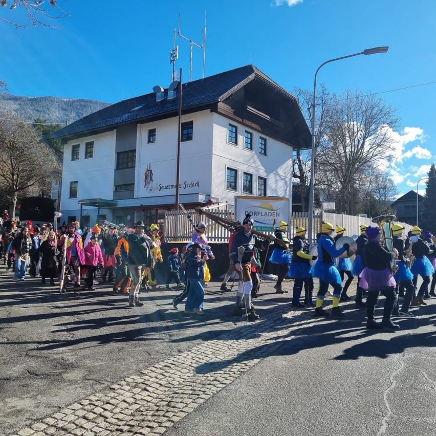 Eine große Gruppe von Menschen in bunten Kostümen marschiert in einem Umzug auf einer Straße, mit einem Gebäude, das ein Schild mit der Aufschrift 'Sennwalder Freilach' im Hintergrund zeigt.
