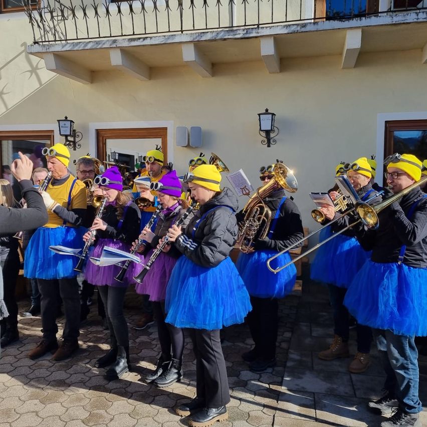 Eine Gruppe von Musikern in bunten Kostümen und Hüten spielt im Freien. Sie spielen verschiedene Instrumente, darunter Trompeten, Posaunen und Flöten. Einige Musiker halten Notenblätter in der Hand. Die Aufführung findet vor einem Gebäude statt.