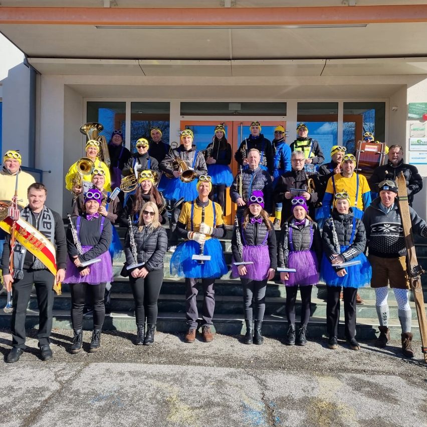 Eine Gruppe von Menschen, die gelbe und lila Tüllröcke tragen, zusammen mit einigen in Skiausrüstung, posiert für ein Foto vor einem Gebäude. Sie halten Musikinstrumente.