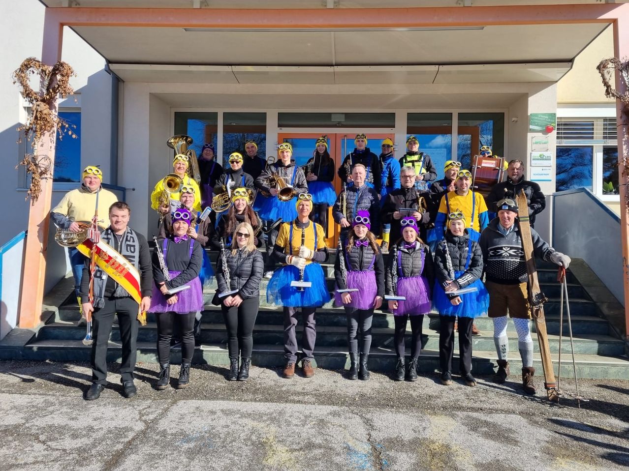 Eine Gruppe von Menschen, die gelbe und lila Tüllröcke tragen, zusammen mit einigen in Skiausrüstung, posiert für ein Foto vor einem Gebäude. Sie halten Musikinstrumente.