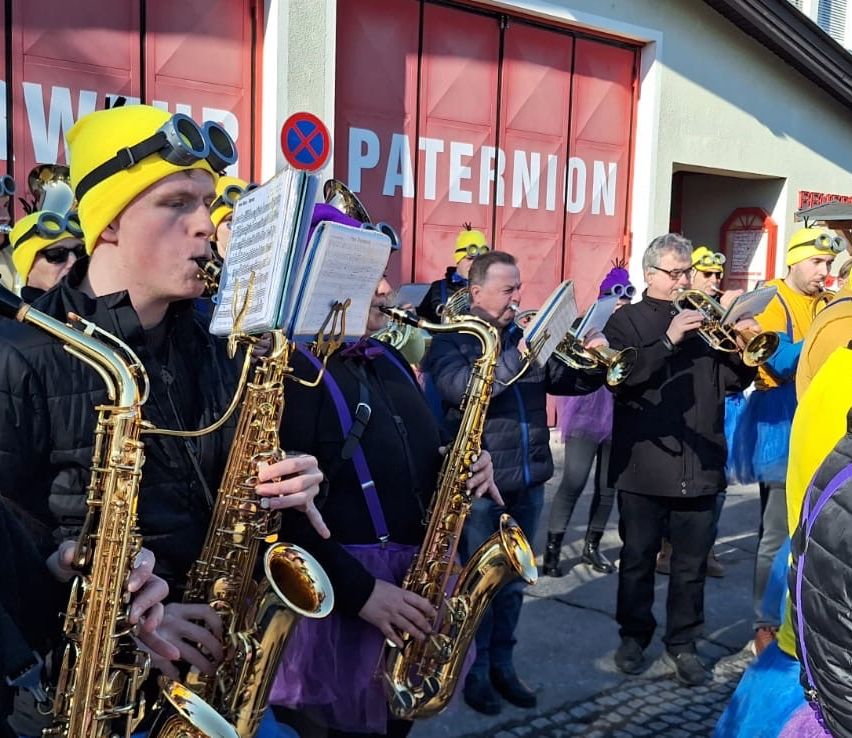 Eine Gruppe von Musikern mit Schutzbrillen und gelben Mützen spielt Saxophone vor einem roten Gebäude mit der Aufschrift 'Paternion'.