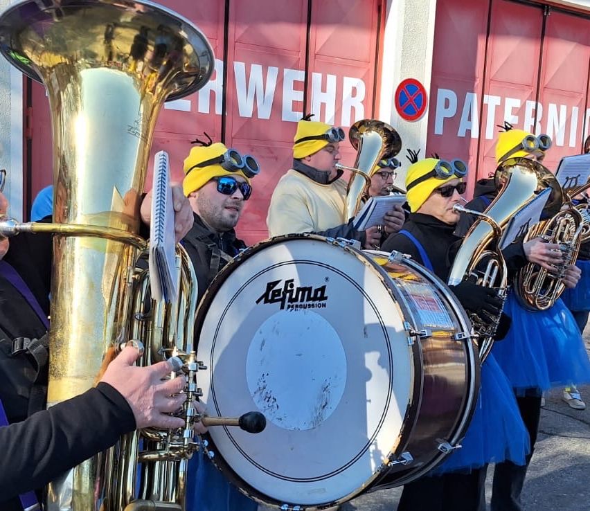 Eine Gruppe von Menschen in blauen Kostümen und gelben Mützen mit Schutzbrillen spielt Musikinstrumente, darunter Tuba und Trommel, vor einem roten Gebäude.