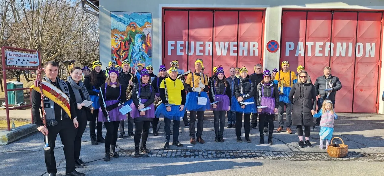 Eine Gruppe von Menschen, die in bunten Tüllröcken und Mützen gekleidet sind, wahrscheinlich Musiker, stehen vor einem Feuerwehrhaus mit einer großen roten Tür. Sie halten Musikinstrumente in den Händen und scheinen für ein Foto zu posieren.