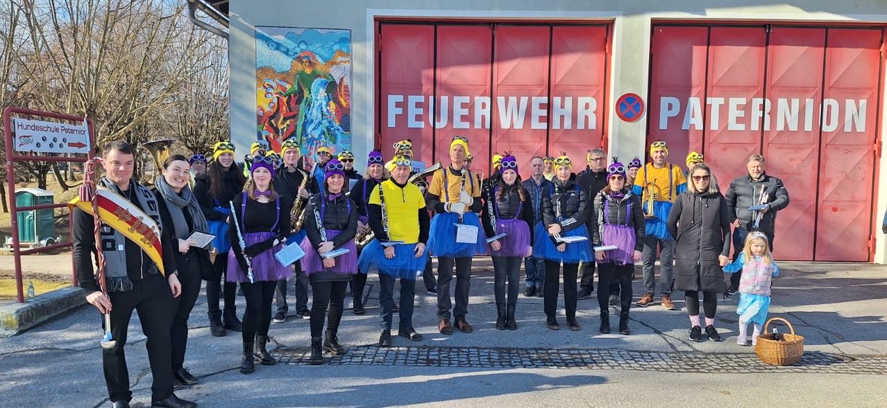 Eine Gruppe von Menschen, die in bunten Tüllröcken und Mützen gekleidet sind, wahrscheinlich Musiker, stehen vor einem Feuerwehrhaus mit einer großen roten Tür. Sie halten Musikinstrumente in den Händen und scheinen für ein Foto zu posieren.