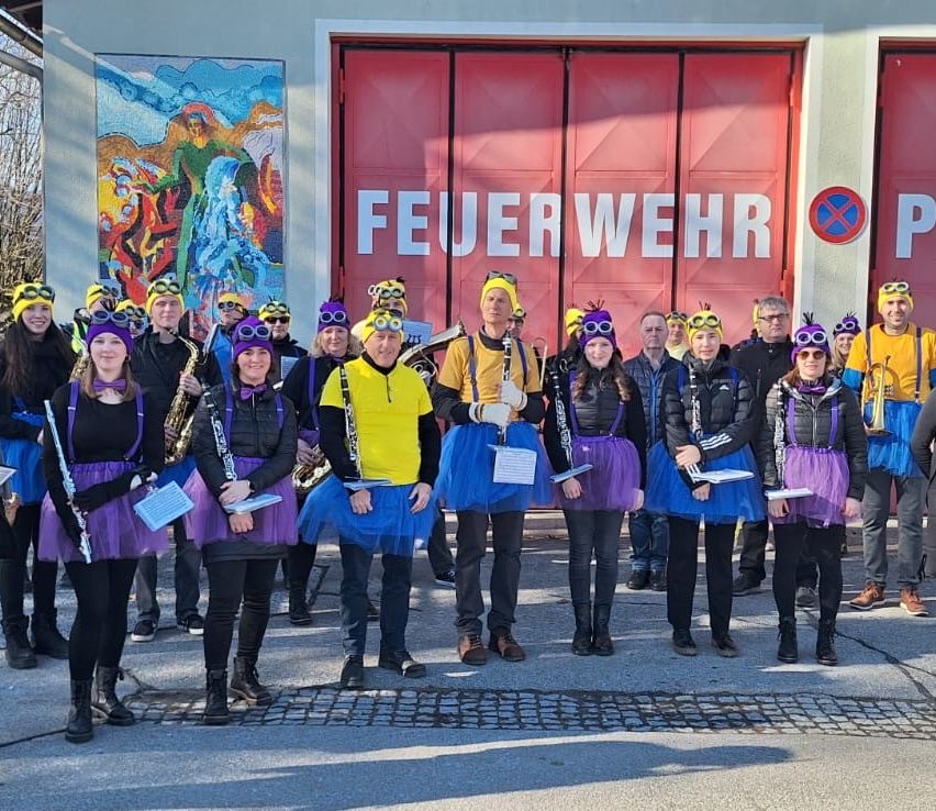 Eine Gruppe von Menschen, die in bunten Tüllröcken und Mützen gekleidet sind, wahrscheinlich Musiker, stehen vor einem Feuerwehrhaus mit einer großen roten Tür. Sie halten Musikinstrumente in den Händen und scheinen für ein Foto zu posieren.