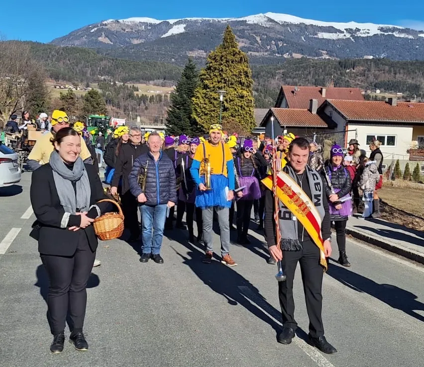 Eine Gruppe von Menschen, einige mit gelben Hüten und Brillen, laufen in einem Umzug auf einer Straße. Dahinter stehen Häuser und Berge.