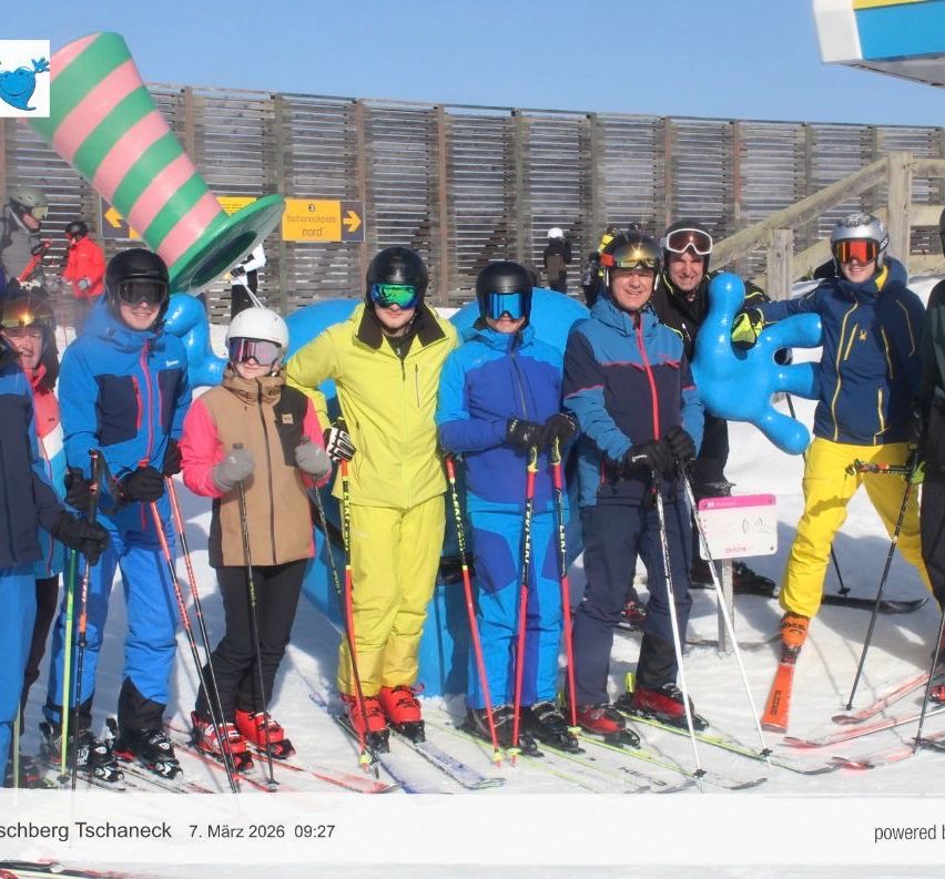Eine Gruppe von Menschen posiert für ein Foto auf einer verschneiten Piste. Sie tragen Skiausrüstung und farbenfrohe Kostüme, einschließlich eines riesigen Hutes. Im Hintergrund sind ein Zaun und ein Schild sichtbar. Datum und Name des Fotografen stehen unten.
