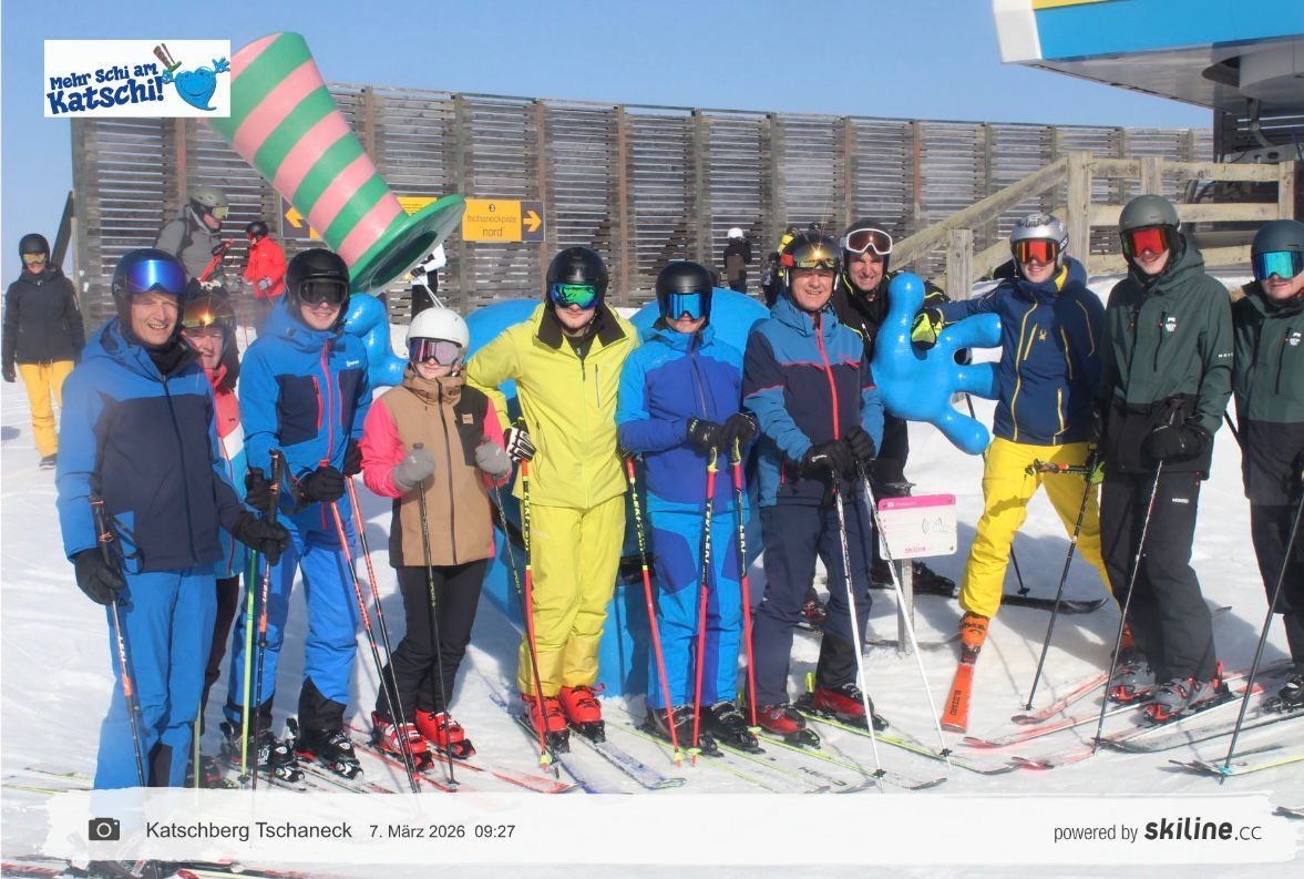 Eine Gruppe von Menschen posiert für ein Foto auf einer verschneiten Piste. Sie tragen Skiausrüstung und farbenfrohe Kostüme, einschließlich eines riesigen Hutes. Im Hintergrund sind ein Zaun und ein Schild sichtbar. Datum und Name des Fotografen stehen unten.