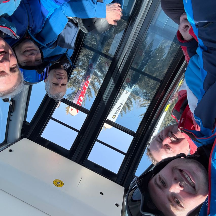 Eine Gruppe von Menschen in Skiausrüstung in einer Gondel, sie lächeln und schauen auf verschneite Bäume.