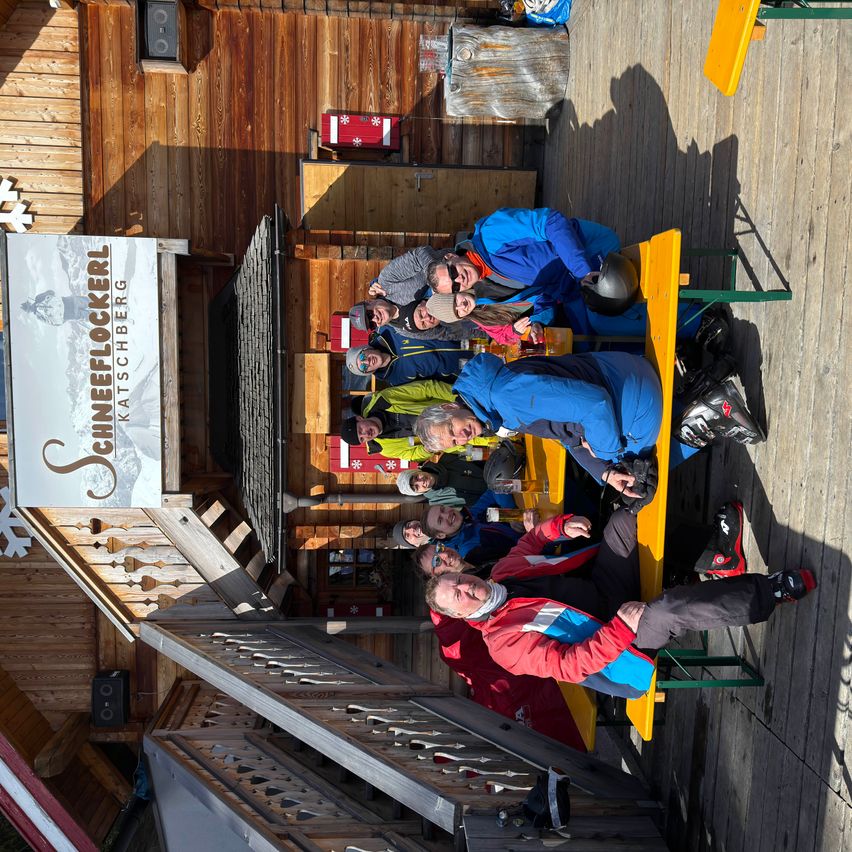 Gruppe von Menschen in Skiausrüstung sitzt auf Bänken vor einer Hütte, ein Schild im Hintergrund trägt den Schriftzug 'Schifleckerl'.