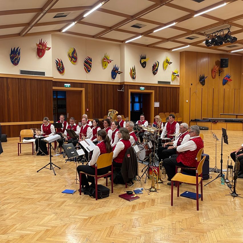 Ein Blechbläserensemble übt in einem Raum mit hölzernen Wänden, die mit farbenfrohen Masken geschmückt sind. Die Musiker sitzen auf Stühlen und spielen verschiedene Instrumente.