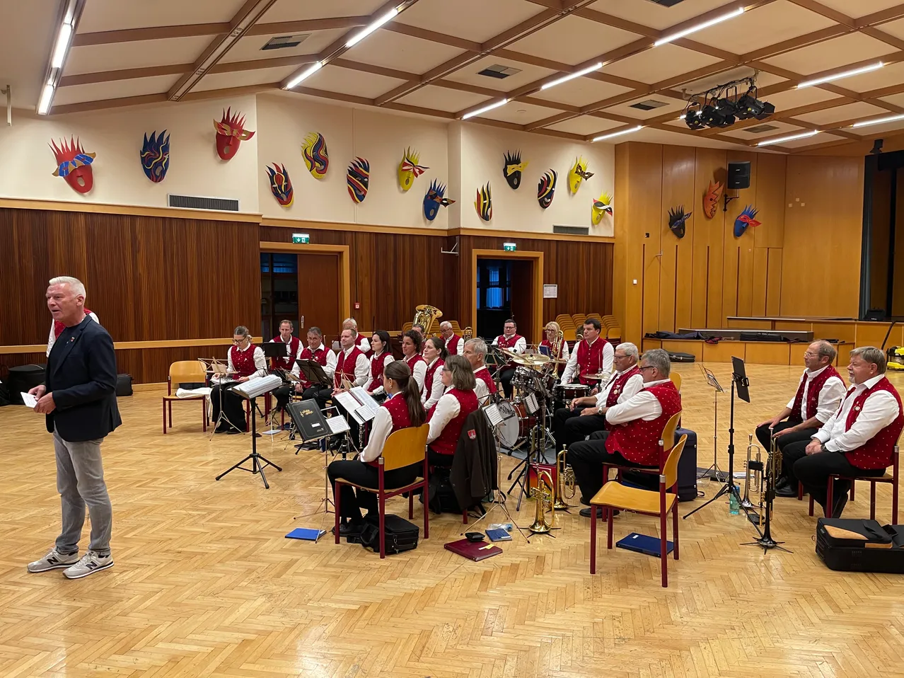 Ein Blechbläserensemble übt in einem Raum mit hölzernen Wänden, die mit farbenfrohen Masken geschmückt sind. Die Musiker sitzen auf Stühlen und spielen verschiedene Instrumente.