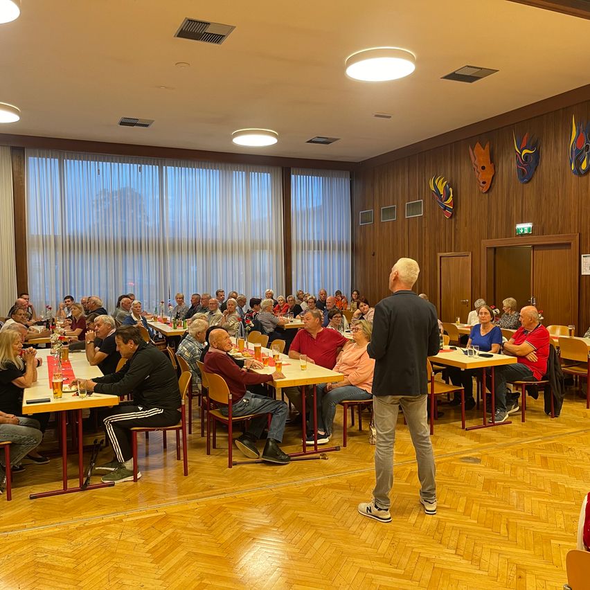 Eine Gruppe von Menschen sitzt an Tischen in einem Raum mit Holzwänden und großen Fenstern. Ein Mann steht vor ihnen und hält möglicherweise eine Rede.