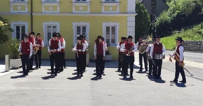 A marching band is performing in front of a yellow building with white trim. They are holding musical instruments.