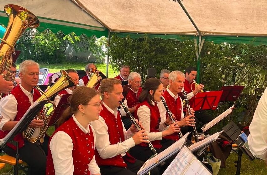 A group of musicians in red vests play instruments under a tent. They include a trumpet, trombone, and clarinet. They are seated with music stands in front of them.